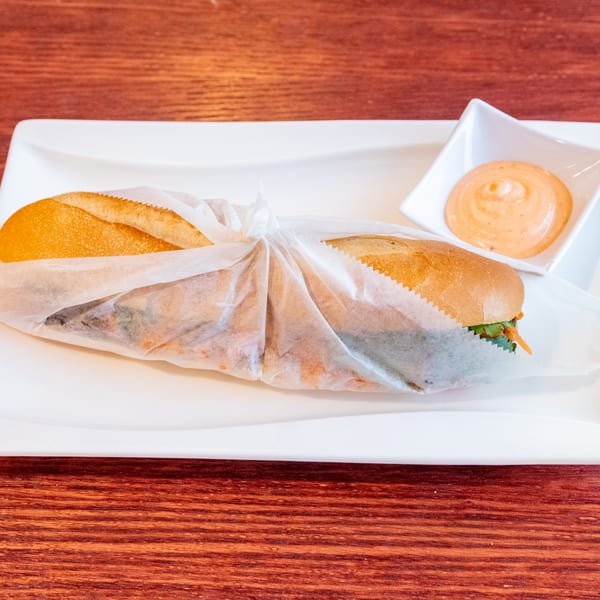 A Vietnamese banh mi sandwich, wrapped in paper, with a side of dipping sauce on a white plate.
