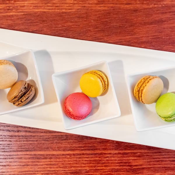 Colorful macarons in small white dishes on a long white tray, against a wooden table.
