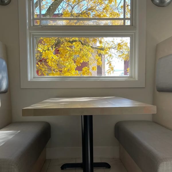 A cozy booth with a light wooden table and benches, looking out a window at bright yellow autumn leaves.